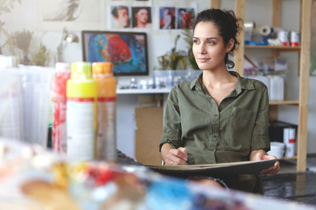 Portrait of young female of creative occupation sitting at modern workshop and working, enjoying process of creating something beautiful, looking sideways with pleased inspired expression on her face